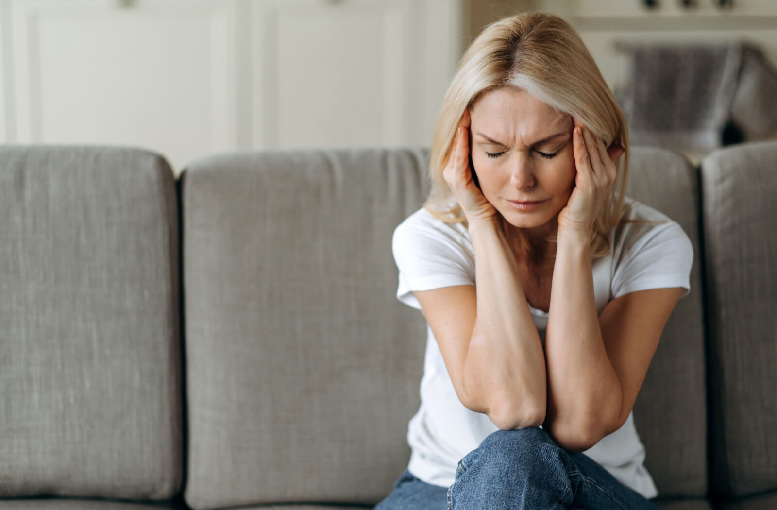 A middle-aged woman sitting on a couch in the living room holding her head with her hands shows fatigue and irritability.