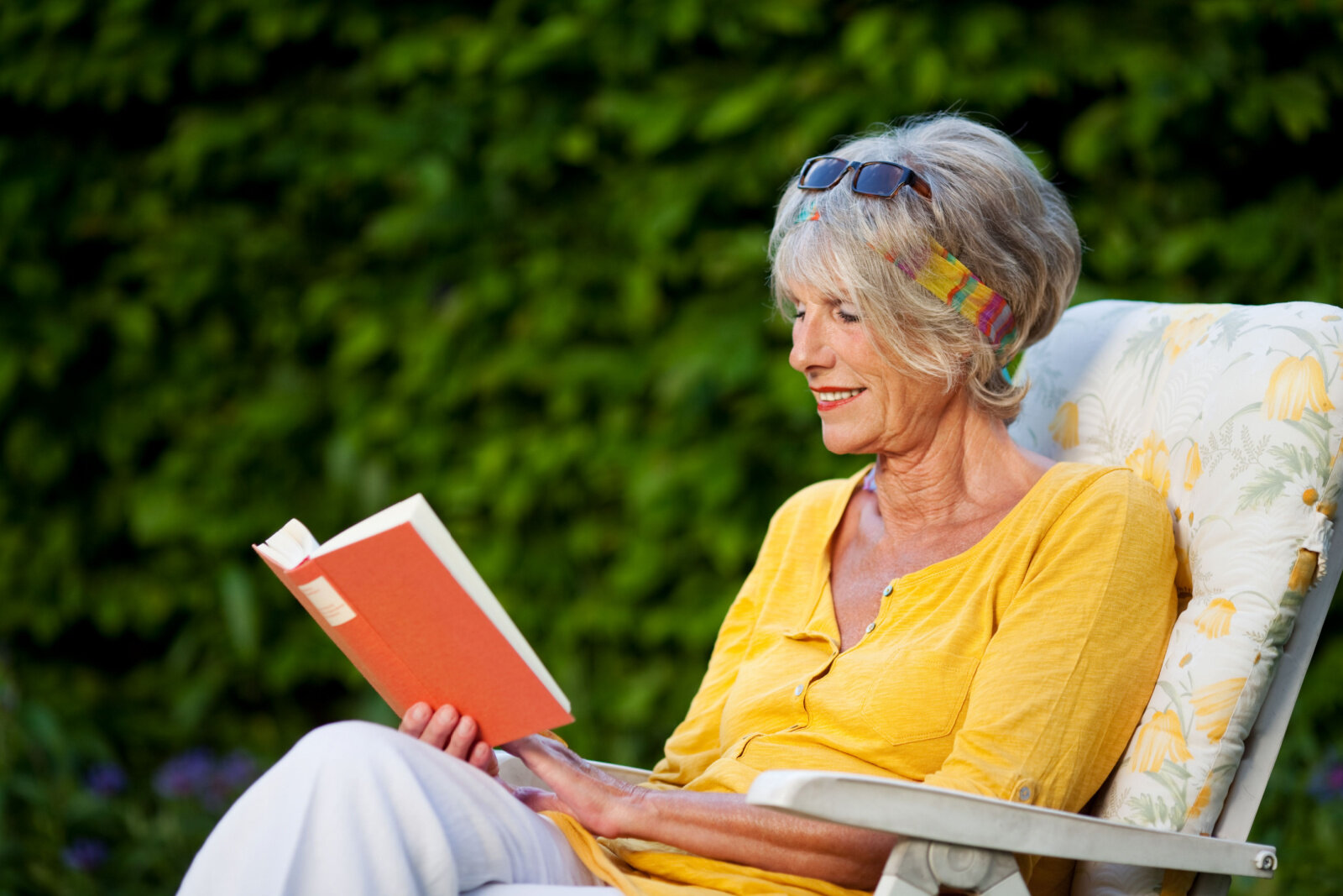 A senior woman is reading a  book while sitting on a chair at the park with a smile on her face.