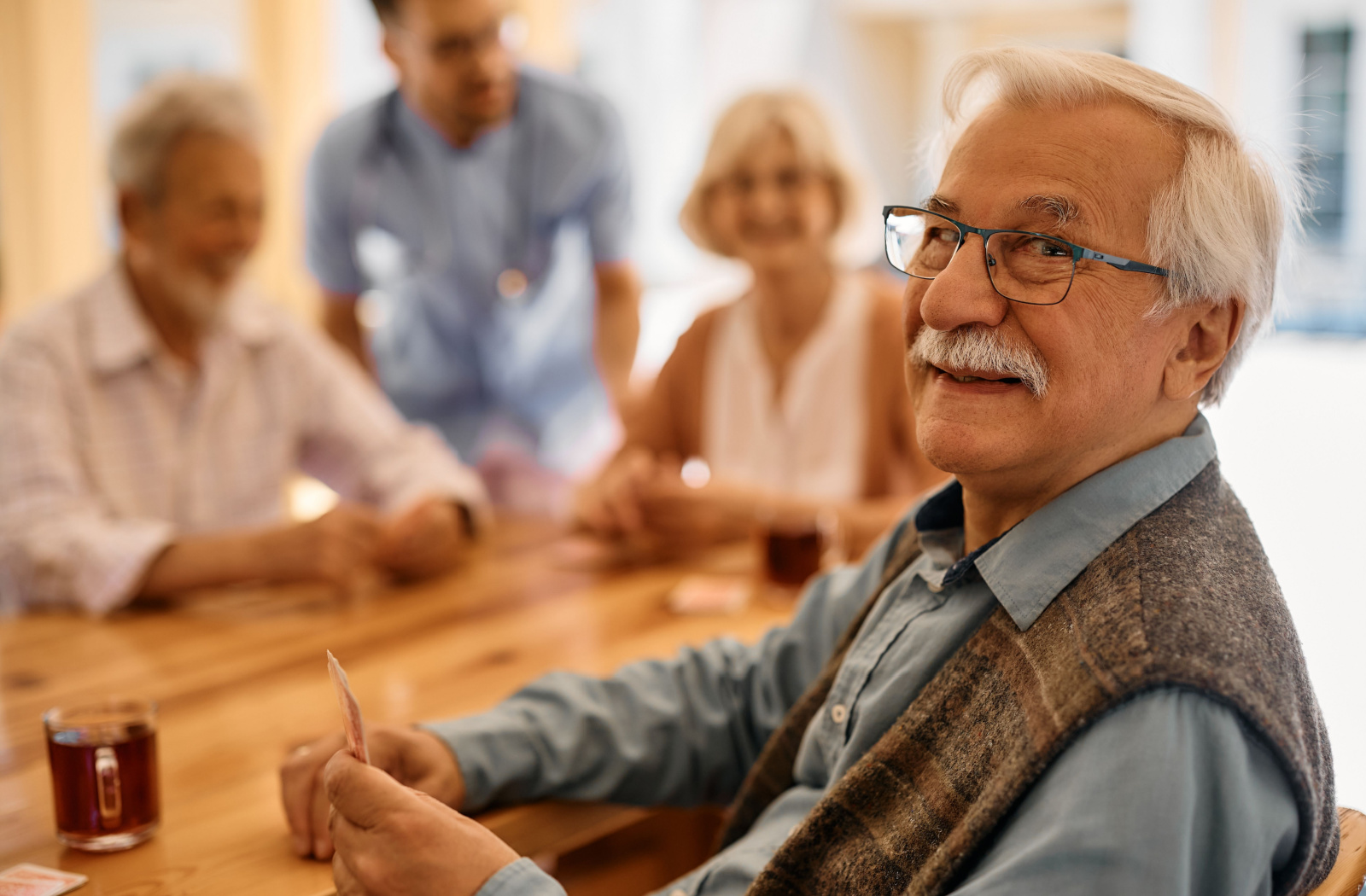 A senior man sits at a table, holding cards next to a cup of tea and smiling. In the background, fellow seniors engage in conversation while a nurse helps out.