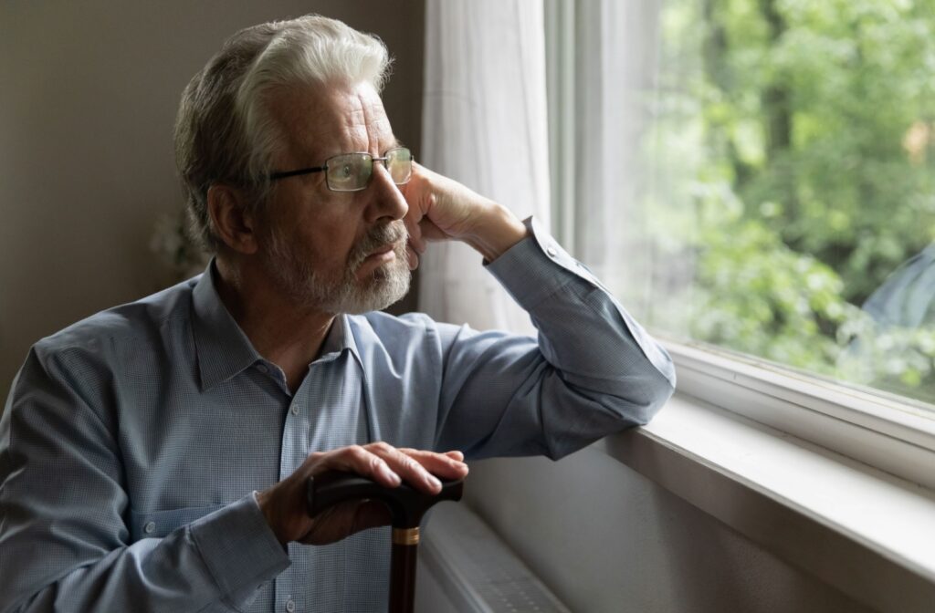 A senior man with glasses leaning on a windowsill and resting his right hand on the top of a cane while looking out the window with a serious expression.