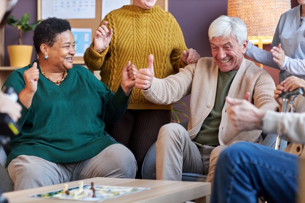 Two seniors celebrate a win in a board game with several friends together in a communal area of a senior living community.