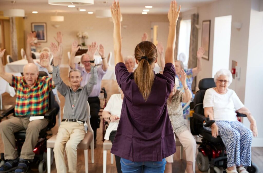 A group of older adults raising their hands while they stay seated as part of a balance exercise class led by a nurse.