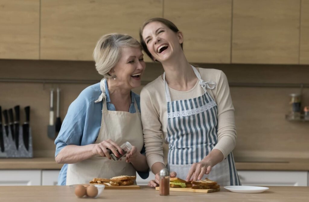 An adult in the kitchen with their senior parent, laughing and making sandwiches together to help address the signs of malnutrition.