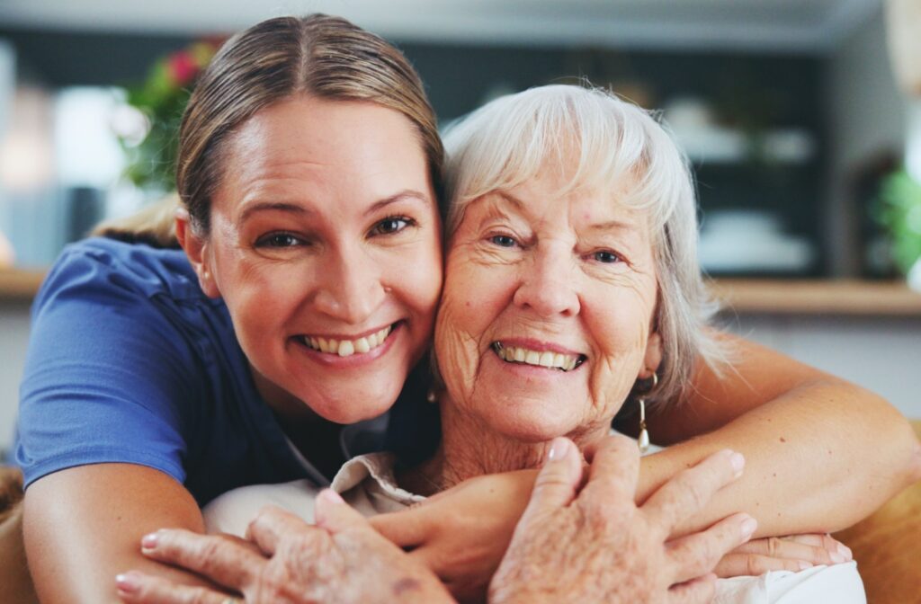 A caregiver hugging an assisted living resident from behind while both smile at the camera.