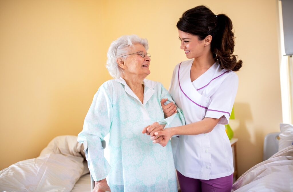 A smiling staff member in assisted living holding an older adult's hand and helping them walk.