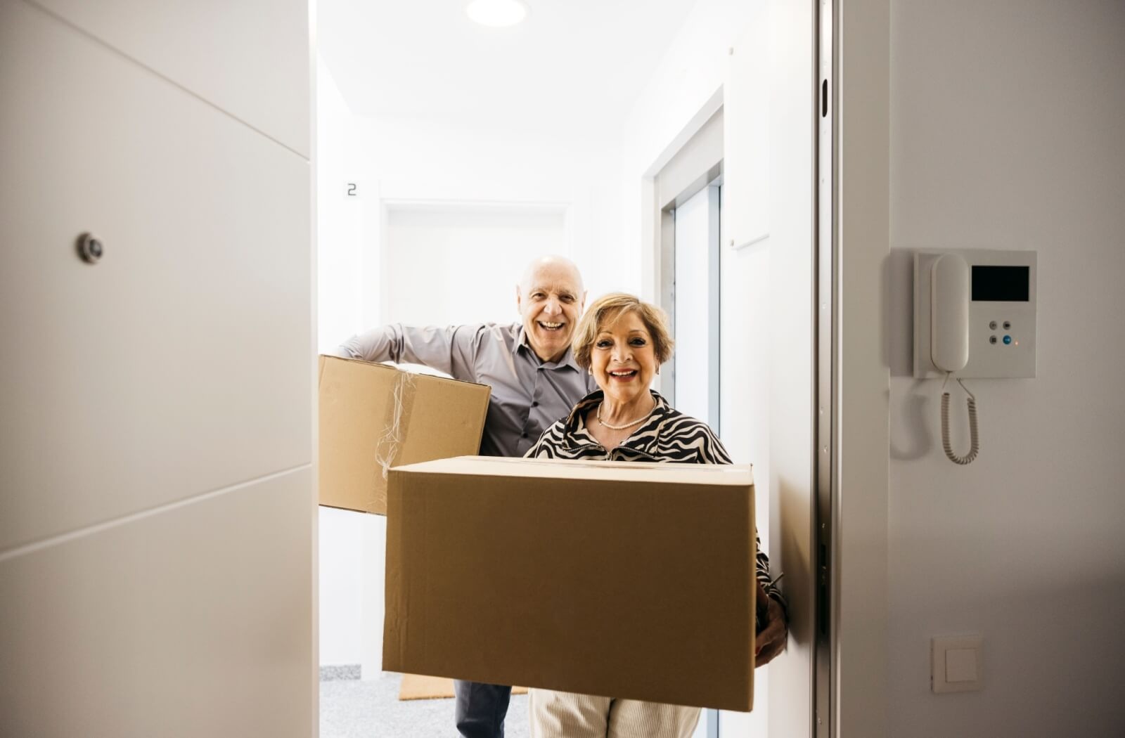 Two older adults carry boxes into a well-it hallway while entering their new home in senior living