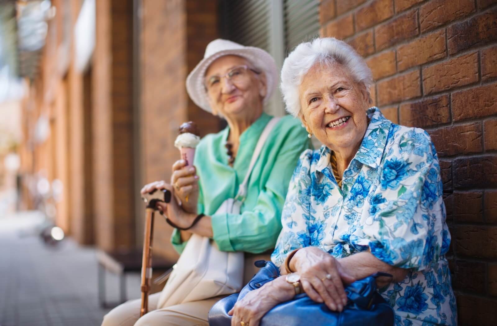 An older adult holding an ice cream cone sits beside their friend and smiles while on a walk outside together
