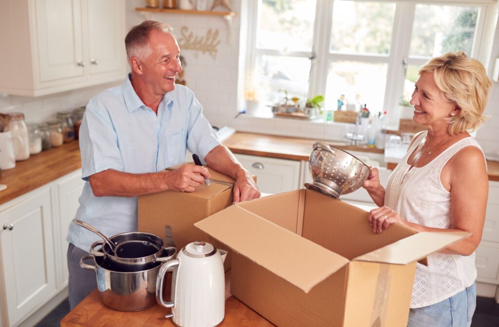 An older adult and their spouse smile at one another while packing dishes and downsizing their kitchen