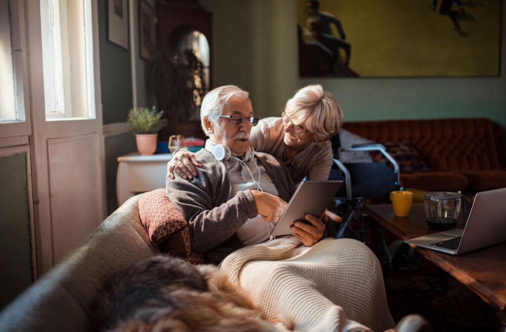 An older adult sitting on their couch at home and showing their spouse pictures of a potential assisted living community on their tablet.