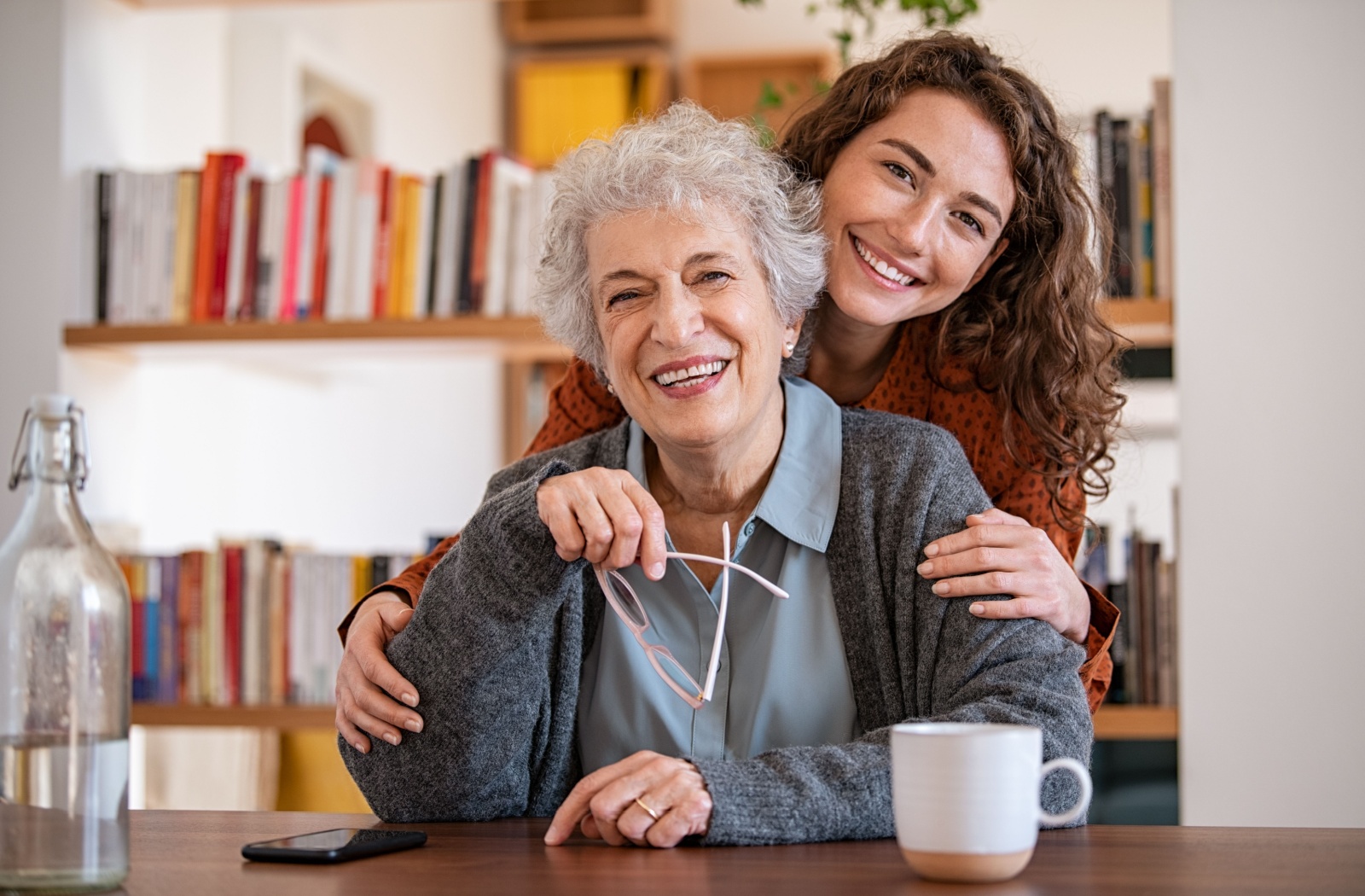 An adult child hugs their older parent from behind, both smiling during a visit to their assisted living home