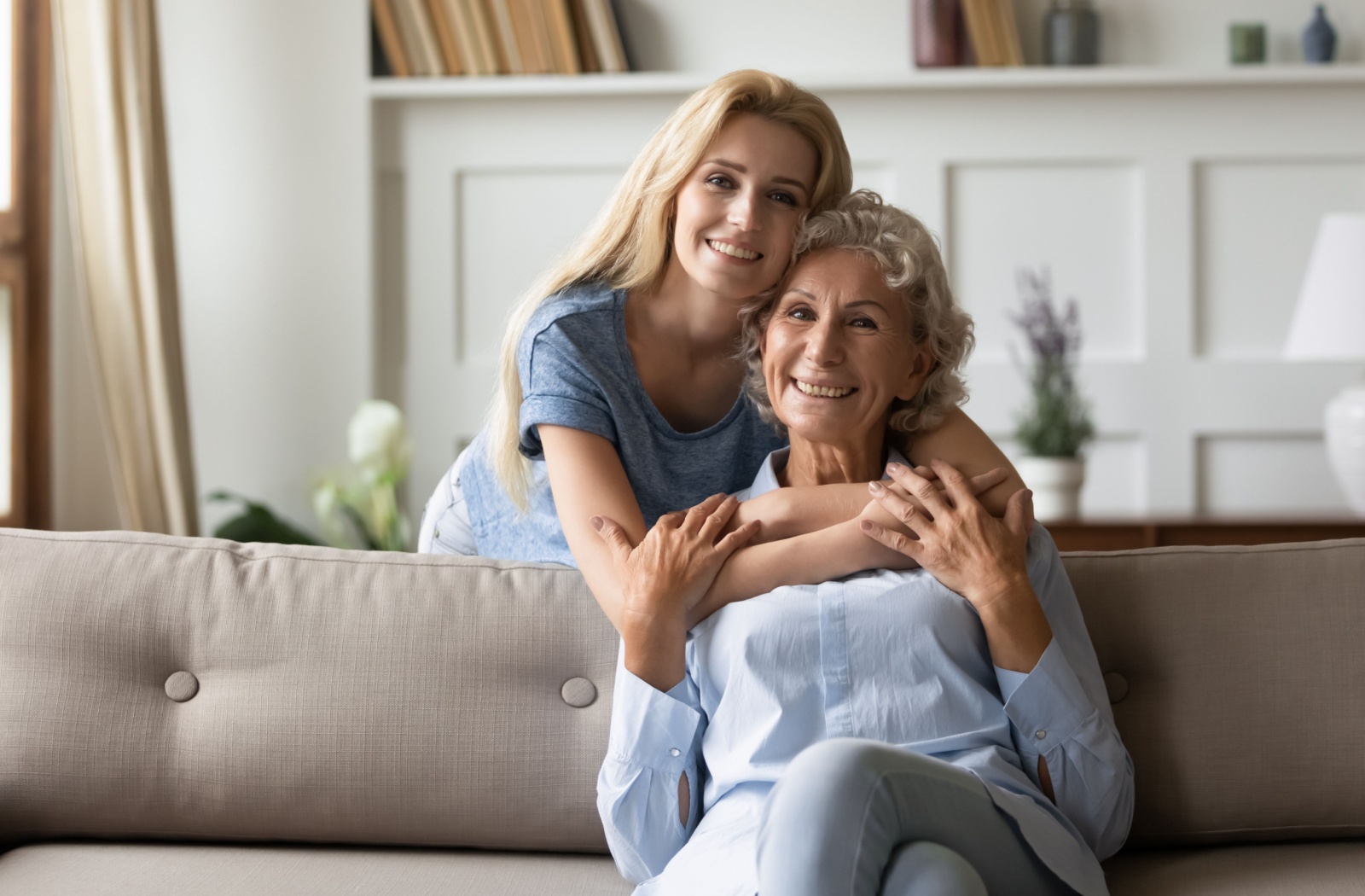 An adult child hugs their senior parent, who is seated on a couch.