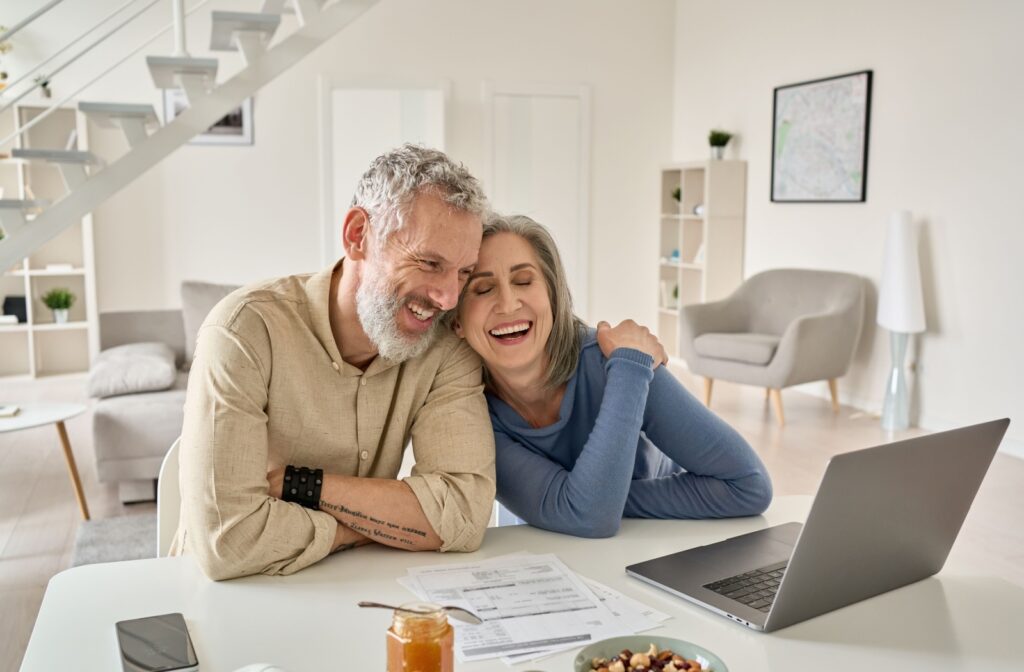 A senior couple leans on each other and smiles as they explore senior living options on a laptop.