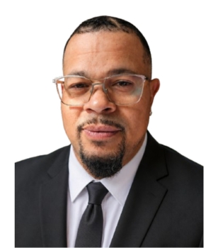 Portrait of a man in a black suit, white shirt, and black tie, wearing glasses.